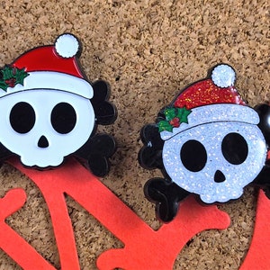 May include: Two enamel pins shaped like skulls with black crossbones behind them. Each skull wears a red Santa hat with a white pom-pom and green holly leaves. One skull has a smooth white finish, while the other has a glittery white finish.