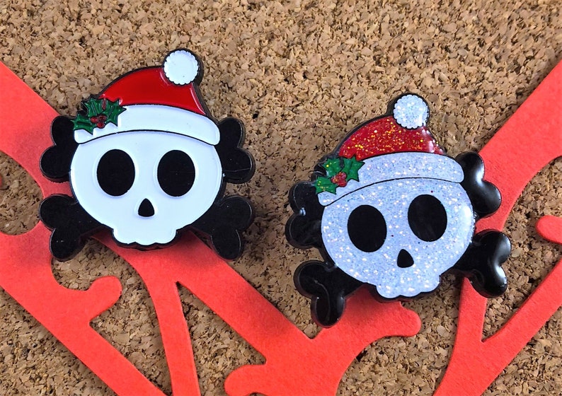 May include: Two enamel pins shaped like skulls with black crossbones behind them. Each skull wears a red Santa hat with a white pom-pom and green holly leaves. One skull has a smooth white finish, while the other has a glittery white finish.
