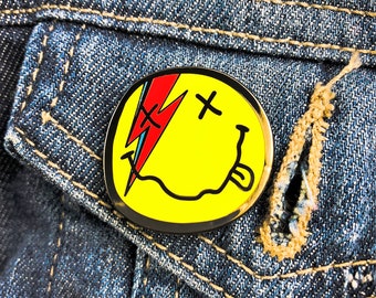 May include: A circular enamel pin featuring a yellow smiley face with a black outline. The face has an 'X' for one eye and a red and blue lightning bolt over the other eye. The mouth is a wavy black line with a small tongue sticking out.