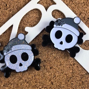 May include: Two enamel pins shaped like skulls with crossbones, each wearing a Santa hat with holly leaves. One skull features a white glitter finish on the face and hat, while the other has a solid white face and a dark grey hat with a white pom-pom.