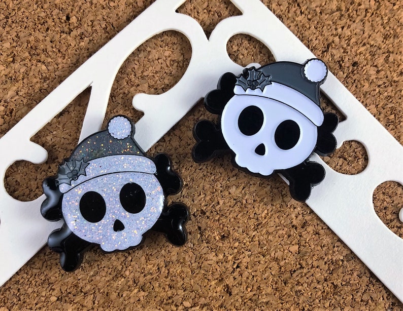 May include: Two enamel pins shaped like skulls with crossbones, each wearing a Santa hat with holly leaves. One skull features a white glitter finish on the face and hat, while the other has a solid white face and a dark grey hat with a white pom-pom.