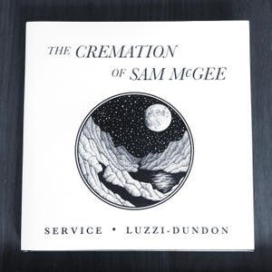 Puede incluir: Una ilustración en blanco y negro de un paisaje iluminado por la luna con montañas y un lago. El texto "THE CREMATION OF SAM McGEE" está en la parte superior de la imagen y "SERVICE • LUZZI-DUNDON" en la parte inferior.
