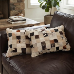 May include: Two square throw pillows featuring a patchwork design made of individual squares of cowhide in various natural shades of white, cream, tan, brown, and black. The pillows have a textured, hairy surface and a visible stitched grid pattern.