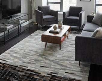 May include: A rectangular area rug featuring a patchwork design of rectangular hide pieces in shades of gray, white, beige, and dark brown. The pattern transitions from a lighter, multi-tonal gray and white center to a dark, banded border.