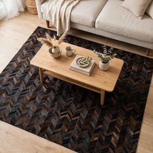May include: A rectangular area rug featuring a dark brown, black, and gray chevron pattern made of stitched panels. The rug is positioned under a light-colored wooden coffee table holding a ceramic bowl, a mug, books, and a small plant.