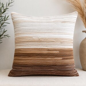 Peut inclure: Un coussin carré présentant un motif dégradé horizontal. Le design passe d'une couleur crème claire en haut, à travers des nuances de beige et de brun clair au milieu, jusqu'à une couleur brun foncé en bas, avec une texture côtelée.
