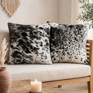 May include: Two square throw pillows featuring a black and white cowhide pattern. The left pillow displays larger, irregular black patches on a white base, while the right pillow showcases a dense, speckled pattern of small black spots on white.
