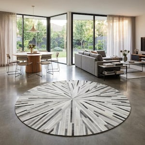 May include: A large circular area rug featuring a sunburst pattern composed of alternating strips of white, grey, and black leather. The rug is positioned on a smooth concrete floor in an open-plan living space with a dining area and sofa.