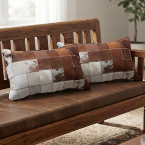 May include: Two rectangular throw pillows featuring a patchwork design made of cowhide squares in varying shades of brown, tan, and white. The pillows have a visible texture and are arranged side by side, showcasing a grid pattern of natural fur.
