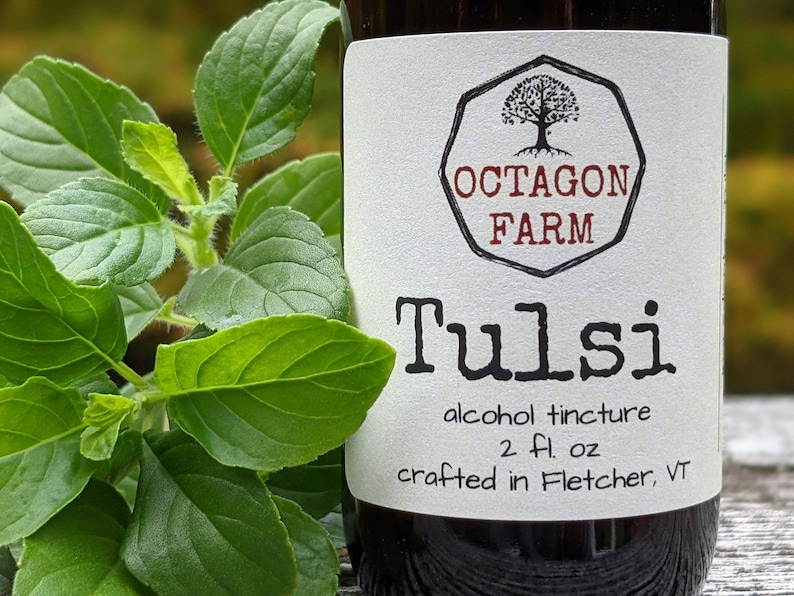 May include: A glass bottle with a white label that reads "Octagon Farm Tulsi alcohol tincture 2 fl. oz crafted in Fletcher, VT". The bottle is sitting next to a green plant.