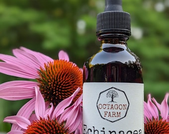 May include: A brown glass bottle of Echinacea alcohol tincture with a black dropper, labeled "Octagon Farm." The bottle is surrounded by pink coneflowers with orange centers. The label reads "2 fl oz crafted in Fletcher, VT."