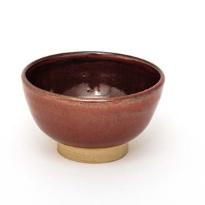Puede incluir: Un cuenco de cerámica rojo con un esmalte marrón y una pequeña base redonda beige.