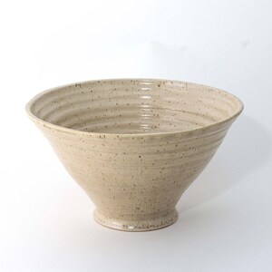 May include: A beige ceramic bowl with a wide, flared shape and a textured surface. The bowl has a footed base and a speckled appearance, suggesting a handmade or rustic aesthetic. The bowl's diameter is approximately 12 inches.