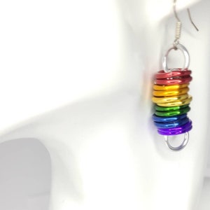 May include: Rainbow coloured metal rings dangle from a silver earring hook. The rings are stacked on top of each other, creating a rainbow effect.