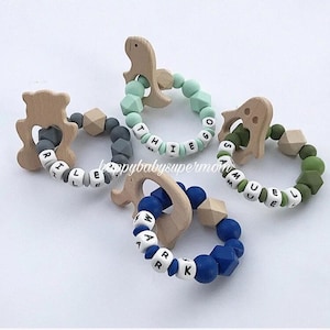May include: Four wooden teething rings with silicone beads and personalized names. The rings are shaped like a dinosaur, a bear, a ghost, and a heart. The beads are in various colors, including green, blue, and gray.
