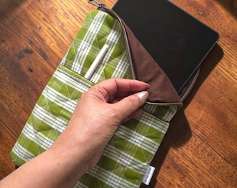 May include: A quilted tablet sleeve featuring a green and white plaid pattern with a brown interior lining and a zipper closure. The sleeve includes an external pocket containing a white stylus and a small white fabric tag on the side.