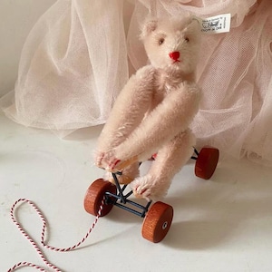 Puede incluir: Un oso de peluche rosa claro montando un triciclo de juguete de madera con ruedas rojas y un marco de metal negro. El triciclo tiene una cuerda atada para tirar de él.