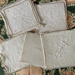 Può includere: Una collezione di quadrati di lino ricamati bianco sporco. Ogni quadrato presenta un disegno diverso, tra cui motivi floreali, una croce e la parola "PAX". I bordi sono rifiniti con pizzo.