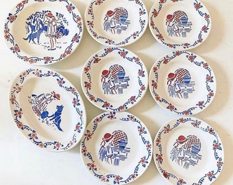 May include: A collection of white ceramic plates with blue and red floral and figure designs. The plates feature a border of blue vines and red flowers. Some plates have a girl with a dog illustration.
