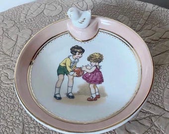 May include: A vintage ceramic ashtray with a pink rim and a gold trim. The ashtray features a painted scene of two children playing with a ball. A white bird figurine is attached to the rim.