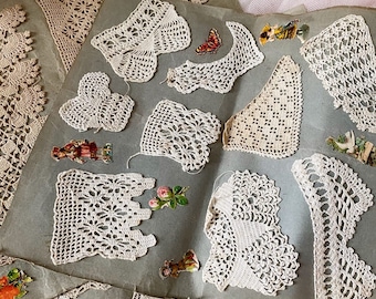 Pode incluir: Uma coleção de peças de renda de crochê antigas, de cor creme, em várias formas e tamanhos, incluindo toalhinhas e bordas. Algumas peças são adornadas com pequenos enfeites de papel coloridos, como borboletas e flores. A renda está disposta sobre uma superfície cinza.