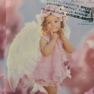 Puede incluir: Una niña pequeña vestida de ángel con alas blancas y plumosas y un vestido rosa. Lleva un sombrero rosa y tiene una dulce expresión en su rostro. El fondo es de un rosa suave con un patrón floral borroso.