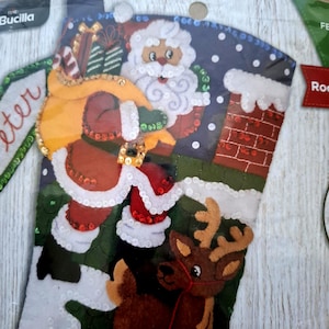 May include: A Bucilla felt Christmas stocking kit featuring a Santa Claus figure carrying a yellow sack of gifts, a reindeer, and a brick chimney. The design is embellished with white and red sequins, with the text Bucilla visible at the top.