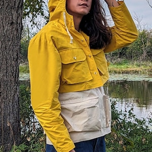 May include: A yellow cropped jacket with a white lining and a hood. The jacket has a zipper and button closure and multiple pockets. The person wearing the jacket is standing in front of a body of water.
