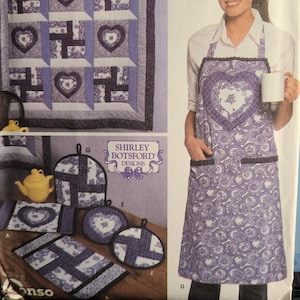 May include: A quilted wall hanging and matching kitchen accessories in shades of purple and white. The quilt features heart and geometric patterns. The apron has a heart design. The accessories include pot holders, a trivet, and a placemat. The brand is Shirley Botsford Designs.