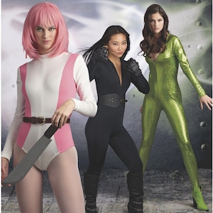 May include: Three women in superhero-themed costumes. One wears a white and pink bodysuit with a brown belt and a toy knife. Another is in a black jumpsuit with a belt and gloves. The third is in a green, textured jumpsuit.