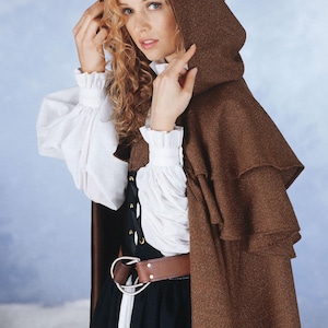 Puede incluir: Una capa con capucha marrón con detalles en capas, usada sobre una blusa blanca con mangas con volantes y un corpiño negro. Un cinturón marrón con hebilla plateada ciñe la cintura. El texto "Simplicity pattern 794" es visible.