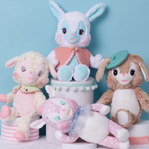 May include: Four plush toys, a white bunny wearing a pink and blue dress, a white lamb wearing a pink dress and green collar, a brown bunny wearing a green beret and a pink and white spotted deer with a blue bow.