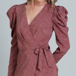 Puede incluir: Un vestido cruzado burdeos con un estampado de puntos blancos. El vestido presenta mangas largas, un escote en V y un lazo en la cintura. Las mangas tienen un diseño de hombros abullonados. El vestido está hecho de una tela ligera.