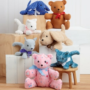 Puede incluir: Una colección de juguetes de peluche, que incluyen un perro azul, un oso marrón, un gato blanco, un gato gris, un perro beige, un oso rosa y un gato azul y blanco. Los juguetes están dispuestos en un estante blanco y una mesa de madera.