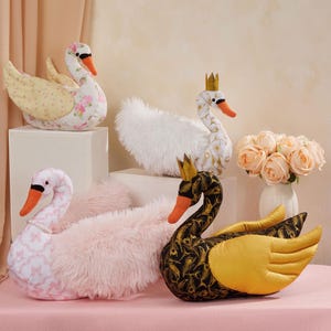 Könnte beinhalten: Eine Sammlung dekorativer Schwan-Plüschtiere in verschiedenen Farben und Mustern. Einer ist weiß mit goldener Krone, ein anderer schwarz mit goldenen Flügeln, weitere mit Blumen- und rosa Designs. Ein Strauß Pfirsichrosen im Hintergrund.