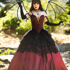 Pode incluir: Uma mulher vestindo um vestido vermelho escuro e preto com uma sobreposição de renda e uma coroa. Ela está segurando grandes asas de morcego pretas.