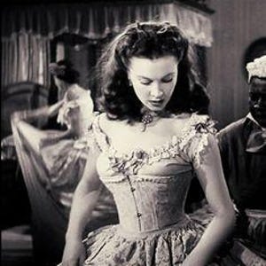Puede incluir: Imagen en blanco y negro de una mujer con un vestido vintage con corpiño de corsé y falda con volantes. El vestido tiene un cuello alto y detalles de encaje delicados. La mujer está mirando hacia abajo, y hay otras figuras en el fondo.