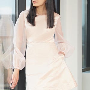 Puede incluir: Un vestido rosa pálido con mangas largas y transparentes y cuello redondo. El vestido tiene un corpiño ajustado y una falda acampanada. Las mangas están recogidas en la muñeca, creando un efecto abullonado. El vestido es de satén brillante.