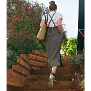 Könnte beinhalten: Eine Person in einem weißen T-Shirt, einem grauen Rock mit Hosenträgern und einer Korbtasche geht eine Holztreppe hinauf. Das Bild enthält den Text "Simplicity Pattern 8641". Die Treppe ist aus Holz und führt zu einem Garten.