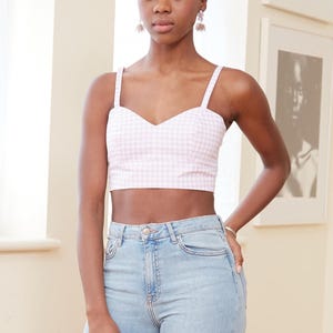 May include: A woman wearing a pink and white gingham crop top with thin straps and light blue jeans.