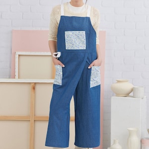Puede incluir: Un delantal de mezclilla azul con un bolsillo de estampado floral blanco y azul. El delantal tiene dos bolsillos grandes en la parte delantera y correas ajustables.