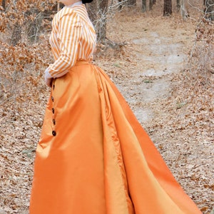 May include: A woman wearing a white and orange striped shirt and a long orange skirt with a bustle. She is wearing a black hat with a feather and is standing in a forest setting.