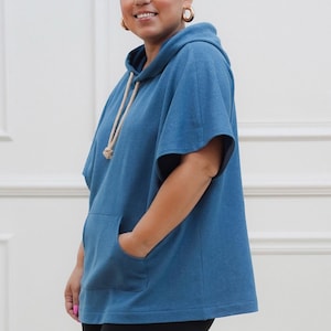 Może przedstawiać: Niebieska bluza z kapturem w stylu poncho z przednią kieszenią i sznurkiem. Top z krótkim rękawem jest wykonany z miękkiego materiału. Modelka ma na sobie czarne legginsy. Tło to biała ściana.