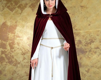 May include: A woman wearing a long, burgundy velvet cape with a white lining and a hood. The cape has a gold clasp at the neck and is worn over a white dress with a gold belt.