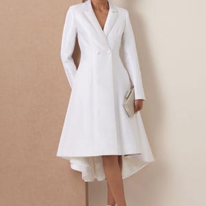 May include: A white coat dress with a double-breasted design and a flared skirt. The dress has long sleeves and a V-neck. The model is holding a silver clutch and wearing silver sandals. The dress is knee-length.