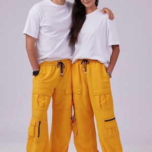 Può includere: Due persone che indossano magliette bianche e pantaloni cargo gialli abbinati con coulisse nere e tasche con cerniera.