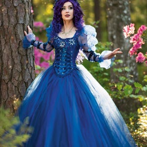 Puede incluir: Una mujer con un vestido de bola azul, una corona floral y cabello morado posa en un entorno de bosque. El vestido tiene un corpiño con cordones y una falda de tul. La mujer está rodeada de árboles verdes y flores rosas.