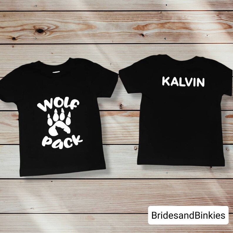 Puede incluir: Dos camisetas negras con texto blanco. Una camiseta dice "Wolf Pack" con una huella de pata. La otra camiseta dice "Kalvin".