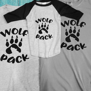 Puede incluir: Tres camisetas grises con letras negras y un gr&aacute;fico de huella de pata. Las camisetas dicen "Wolf Pack".