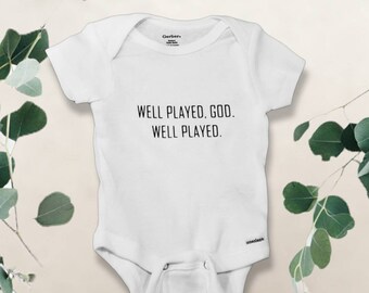 May include: White baby onesie with the text "WELL PLAYED, GOD. WELL PLAYED." in black. The onesie has short sleeves and a snap closure. The brand name "Gerber" is visible on the top of the onesie.